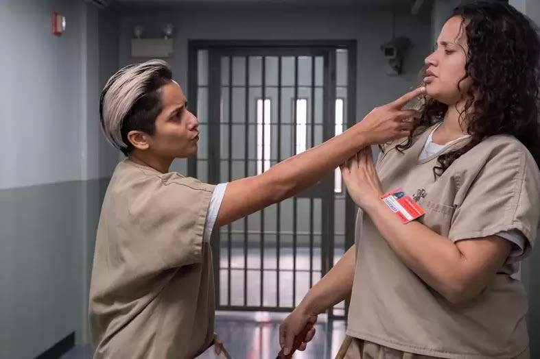 Two women in prison jumpsuits, the women on the left extends her arm towards the women on the right in a threatening manner.